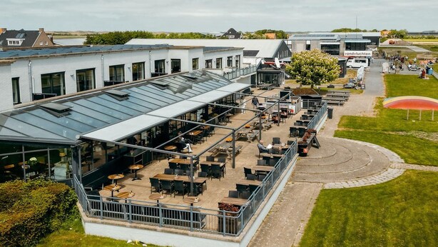 An outdoor cafe area with tables and chairs, a glass-roofed structure, and a grassy lawn.