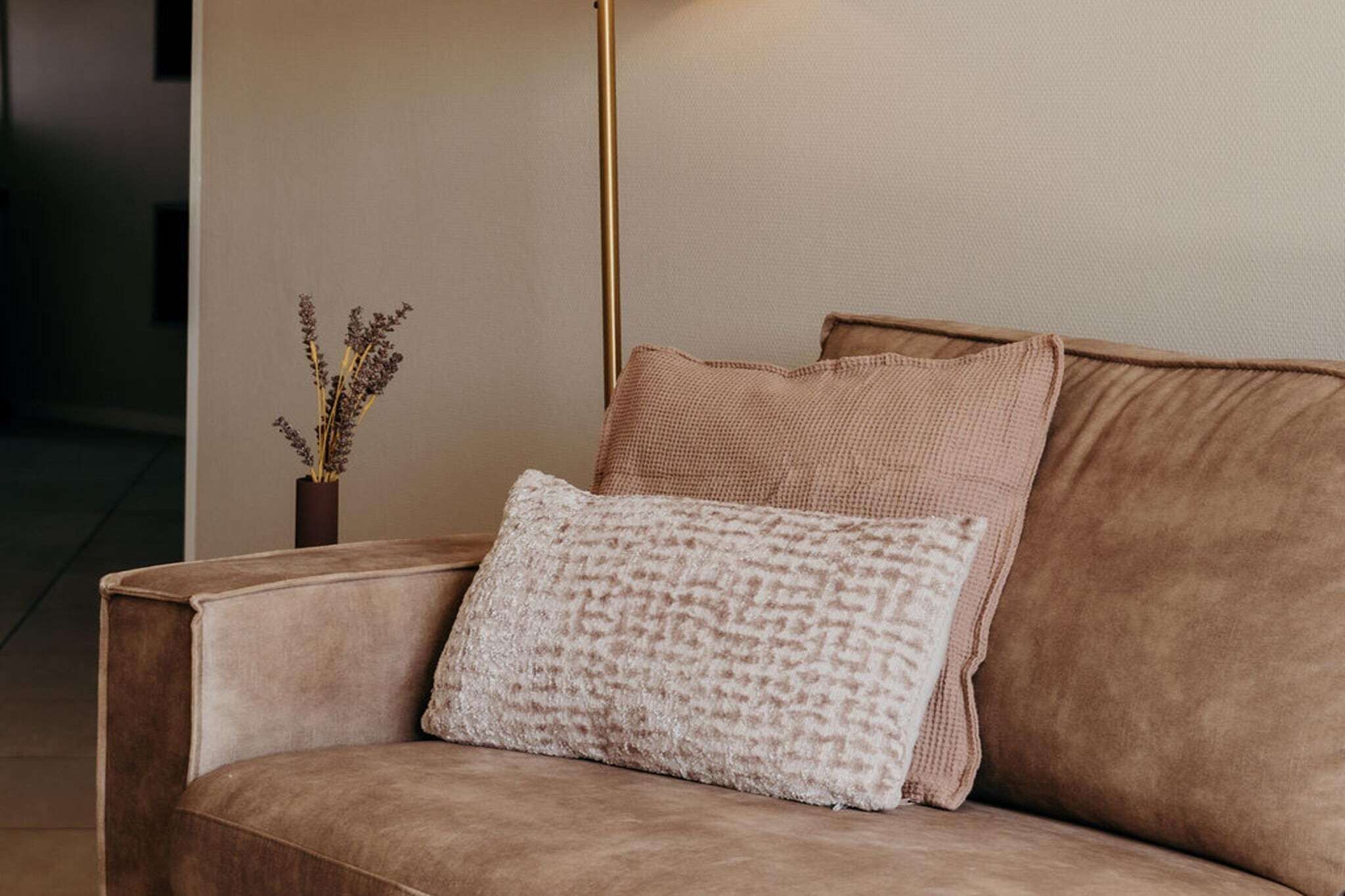 Un sofá marrón con dos almohadas, una blanca y otra marrón, en una habitación con un jarrón de flores secas sobre una mesa.