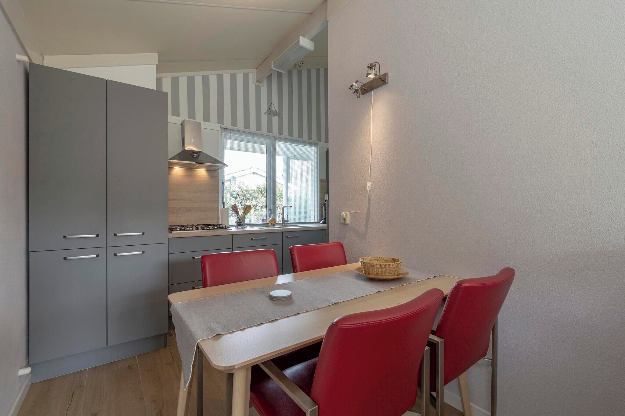A compact kitchen with grey cabinets, a dining table with red chairs, and a window with blinds.