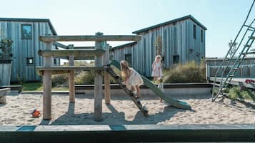 Une structure de jeu en bois avec un toboggan, située dans un espace sablonneux, avec des bâtiments en arrière-plan.
