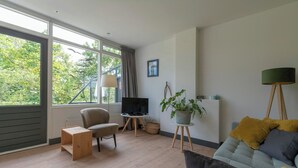 A modern living room with a sofa, a chair, a TV on a stand, a potted plant, and a floor lamp. - De Zeester 4-person holiday home n De Koog (Callantsoog)