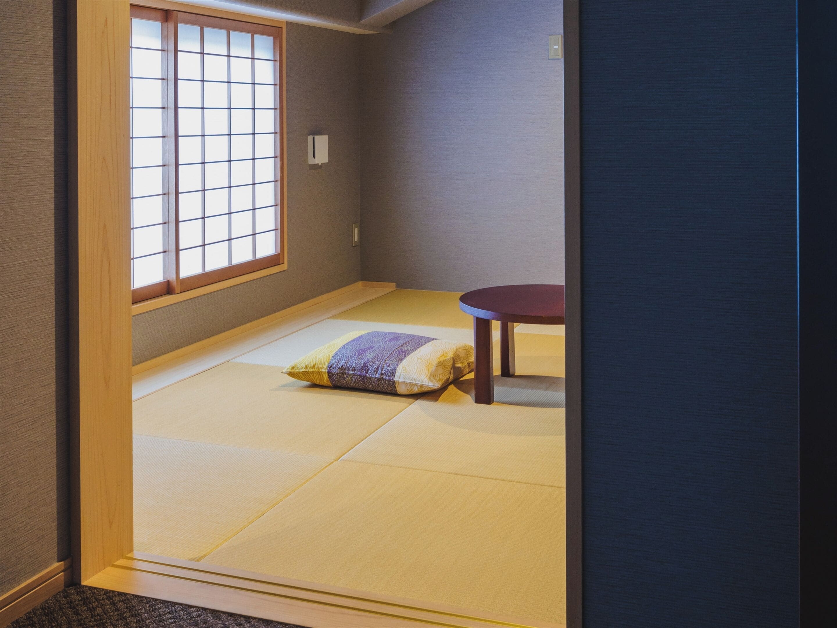 A tatami mat room with a sliding shoji door, a tatami mat, a cushion, and a small round table.