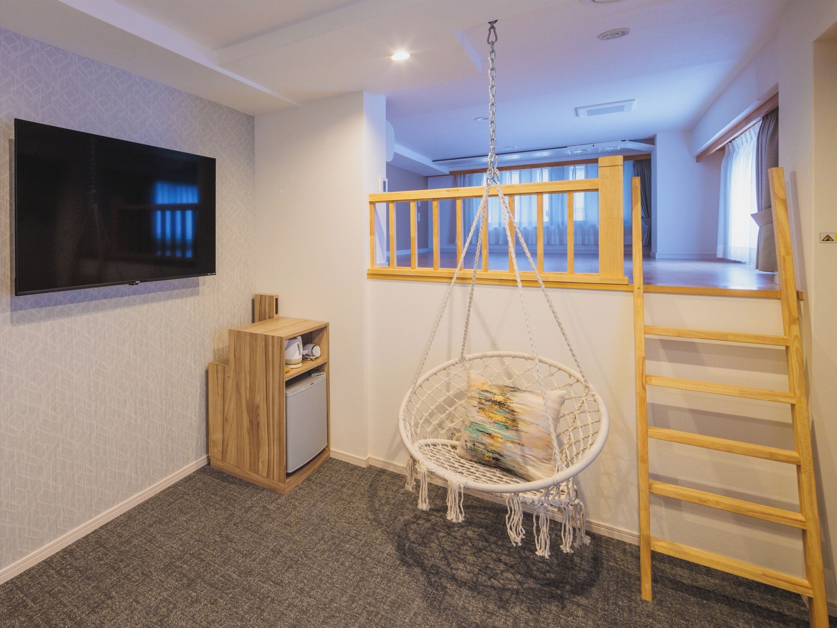 A room with a TV mounted on the wall, a wooden cabinet, a hanging chair, a ladder, and a bunk bed with a wooden railing.