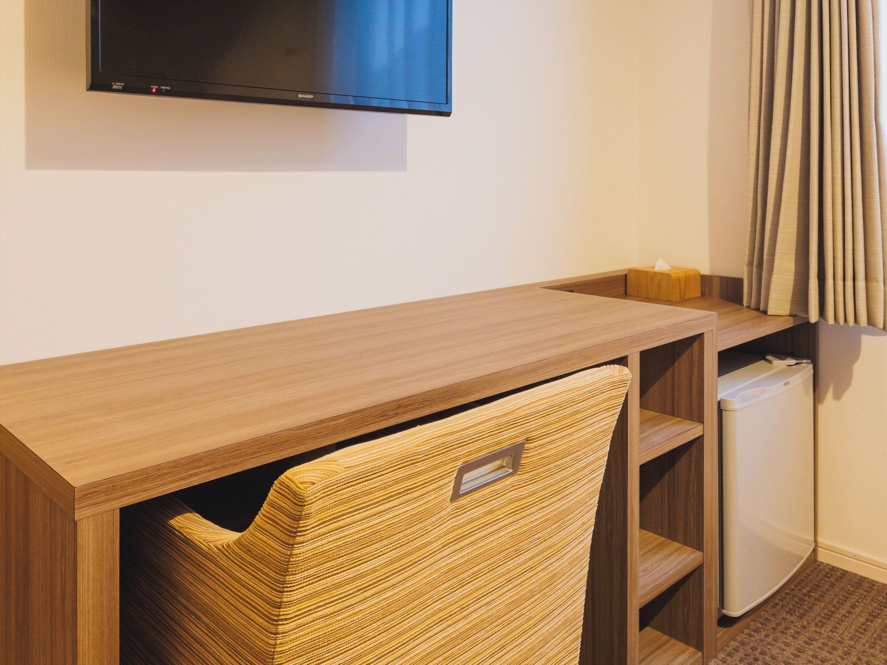 A hotel room with a wooden desk, a television mounted on the wall, a small refrigerator, and a window with curtains.