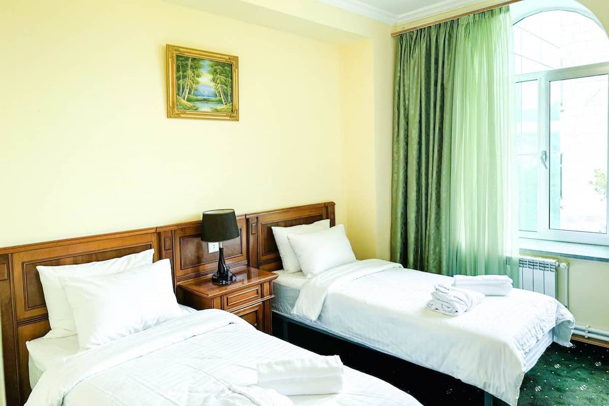 A hotel room with two beds, a nightstand with a lamp, a painting on the wall, and a window with green curtains.