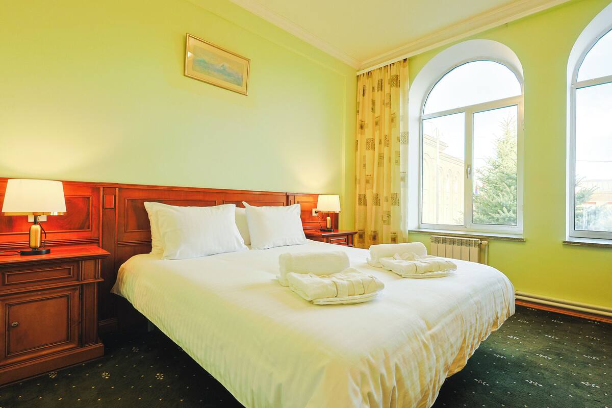 A hotel room with a large bed, two bedside tables with lamps, a window with curtains, and a painting on the wall.