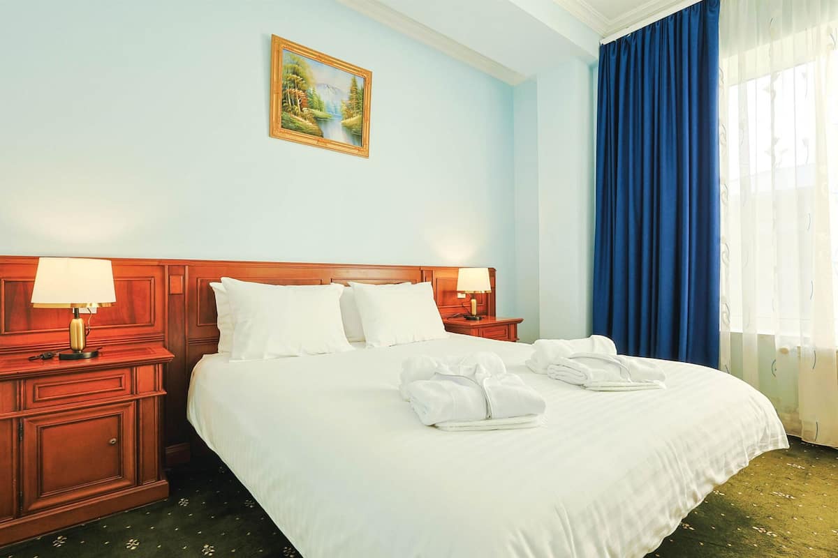 A hotel room with a large bed, two bedside tables with lamps, a painting on the wall, and a window with blue curtains.