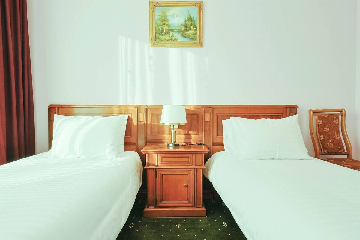 A hotel room with two beds, a nightstand with a lamp, a painting on the wall, and a window with curtains.