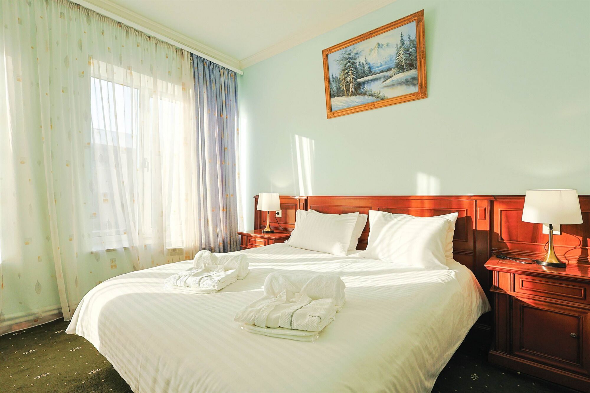 A hotel room with a large bed, two bedside tables with lamps, a painting on the wall, and sheer curtains.
