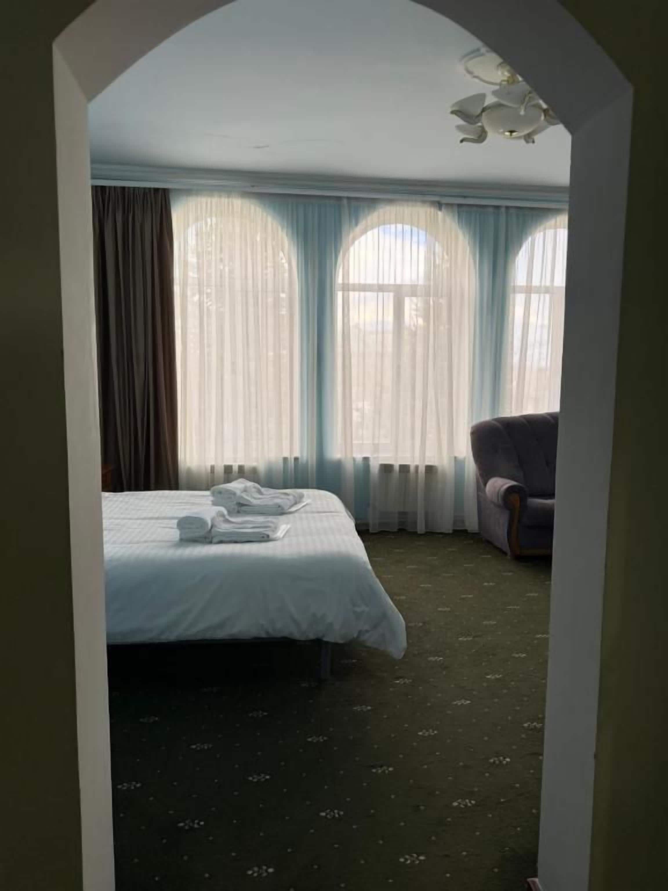 Una habitación de hotel con cama, una silla y ventanas grandes con cortinas.