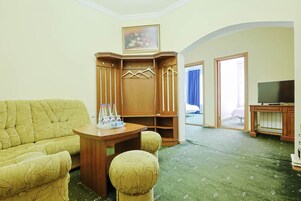 Una habitación de hotel con alfombra verde, un sofá, un armario de madera con ropa colgada, un televisor y un cuadro en la pared.