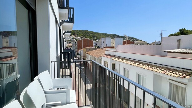 Un balcon avec des chaises blanches, une rambarde en métal et une vue sur les bâtiments voisins et une colline.
