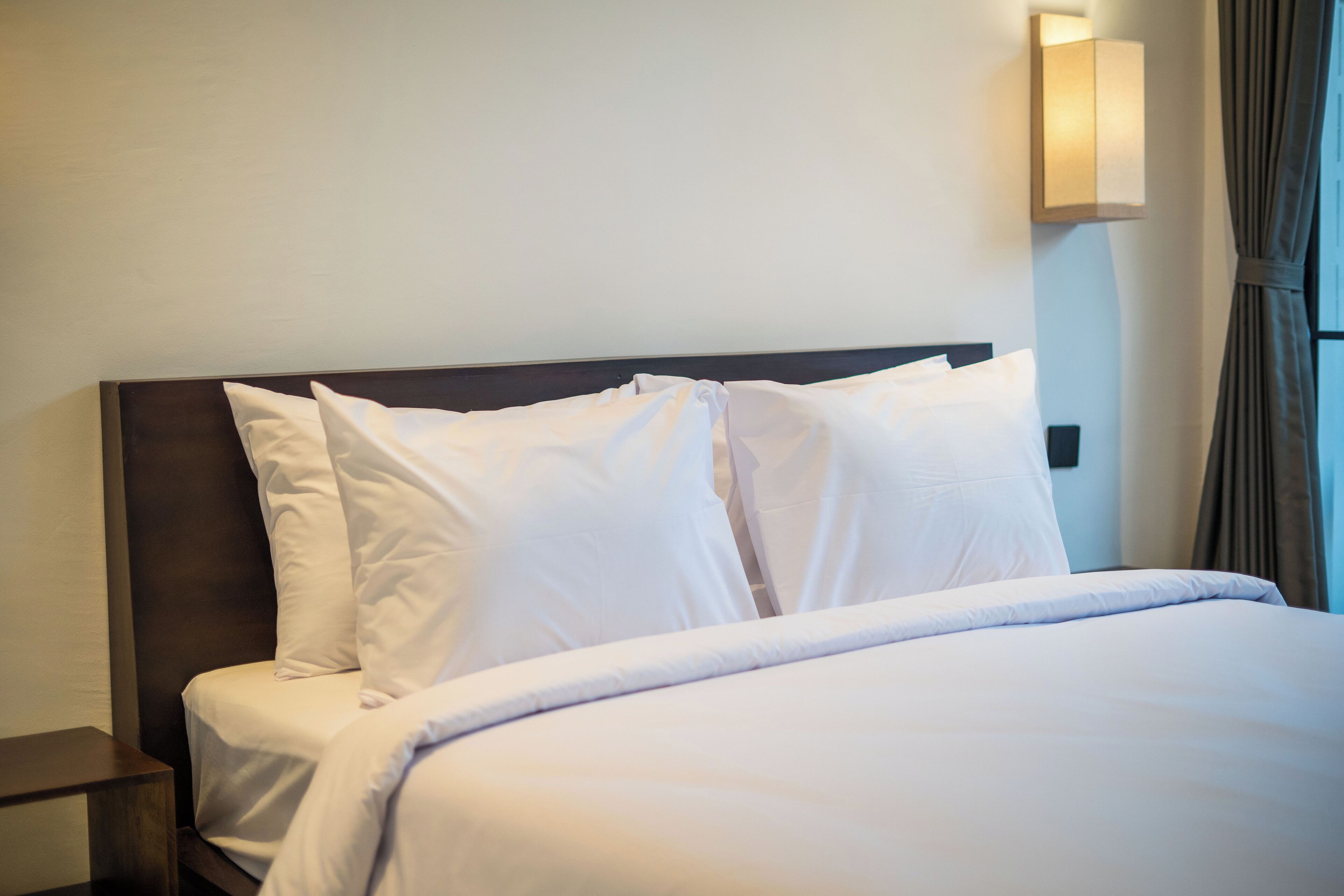 Una cama bien hecha con sábanas y almohadas blancas, un cabecero oscuro y una lámpara de pared.