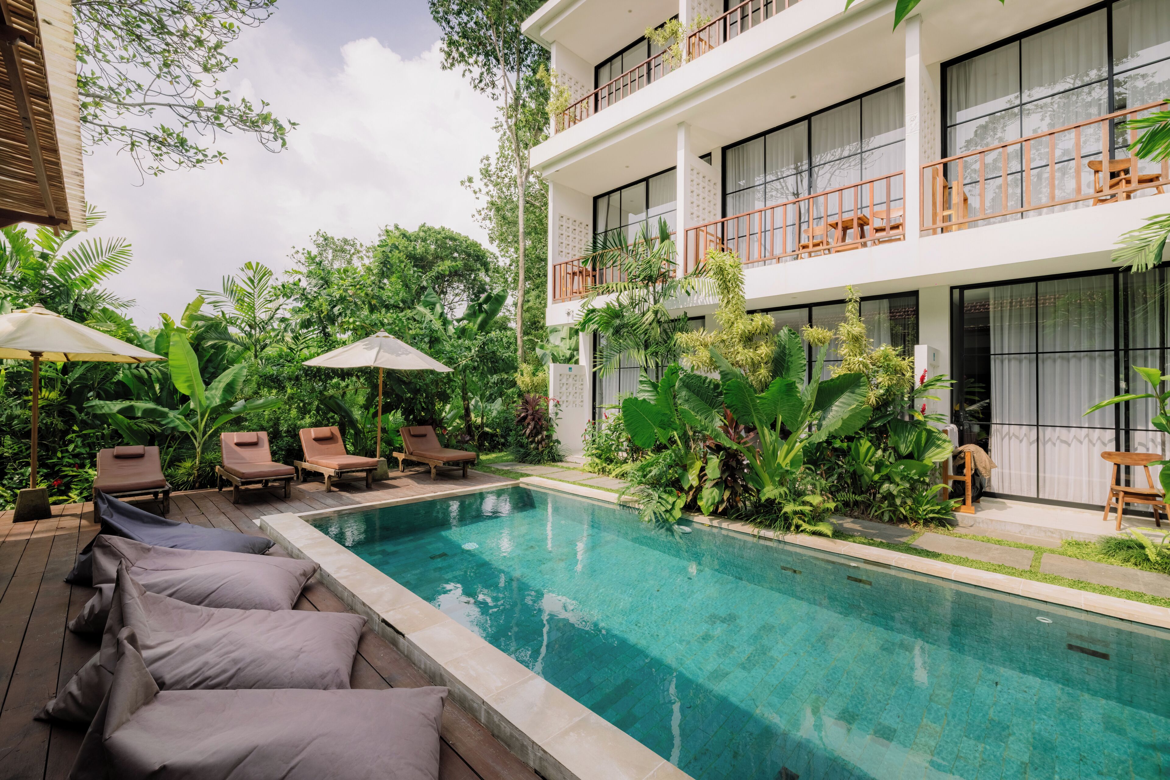 A pool area with lounge chairs and umbrellas, surrounded by lush greenery and a two-story building with balconies.