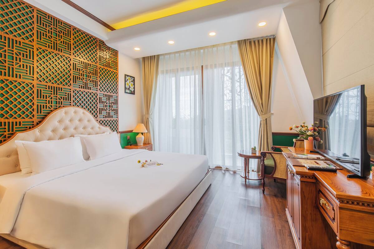 Una cama grande con cabecera capitonada, un escritorio de madera con televisor y un panel decorativo en la pared.