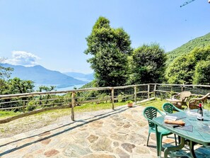 Un patio de piedra con mobiliario exterior, vistas a montañas y un lago, y una baranda de madera.