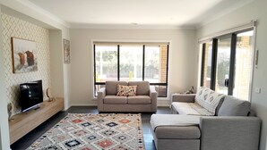 A modern living room with a large window, a grey sofa, a patterned rug, and a flat-screen TV on a wooden stand.