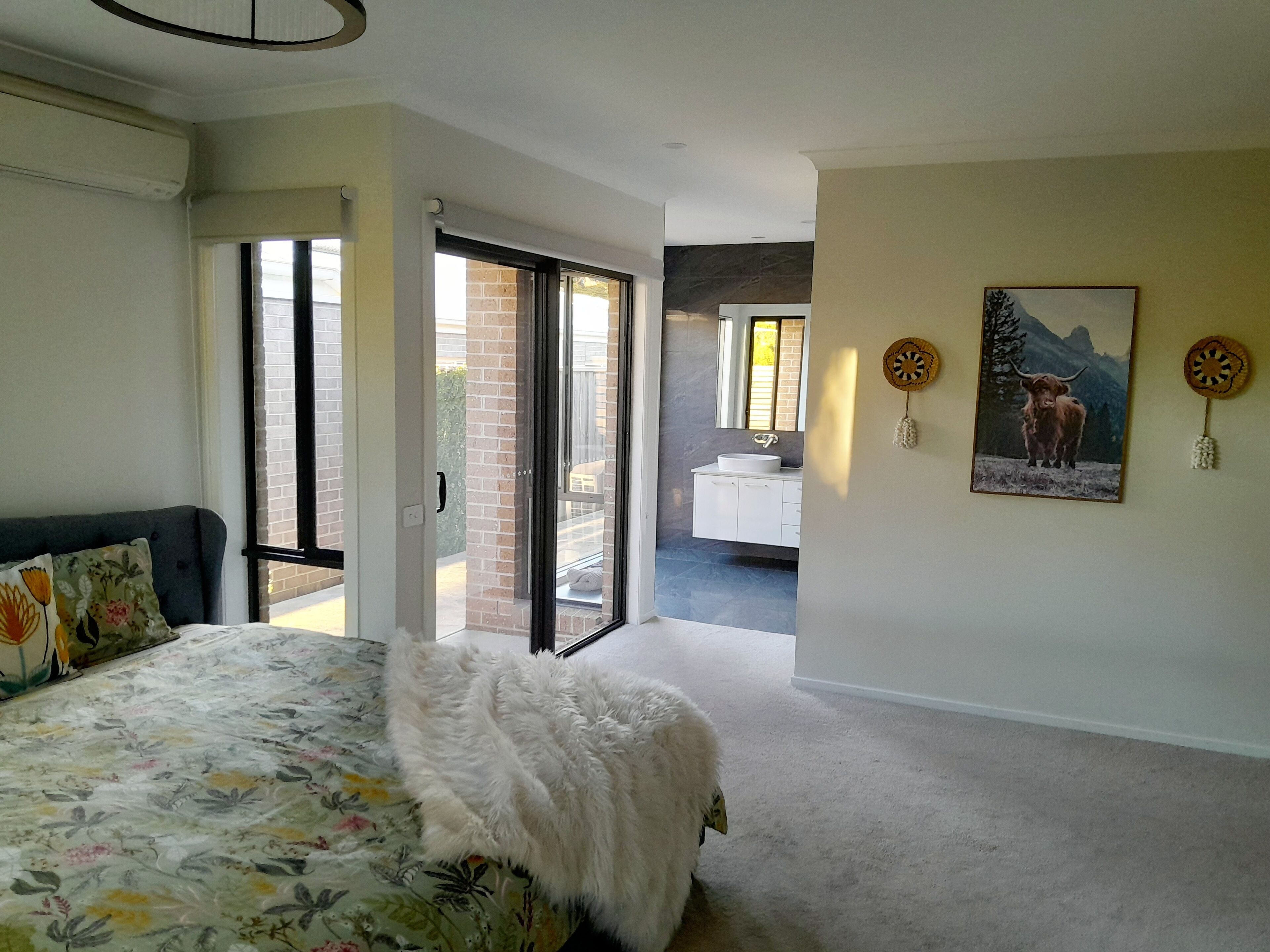A bedroom with a bed, a fluffy white throw, a large window, and a bathroom visible through an open door.