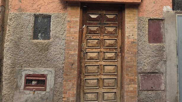 A weathered wooden door with a metal latch, set in a textured wall with a small window and a door frame. - Local Moroccan Style House, Central Medina (Marrakech)