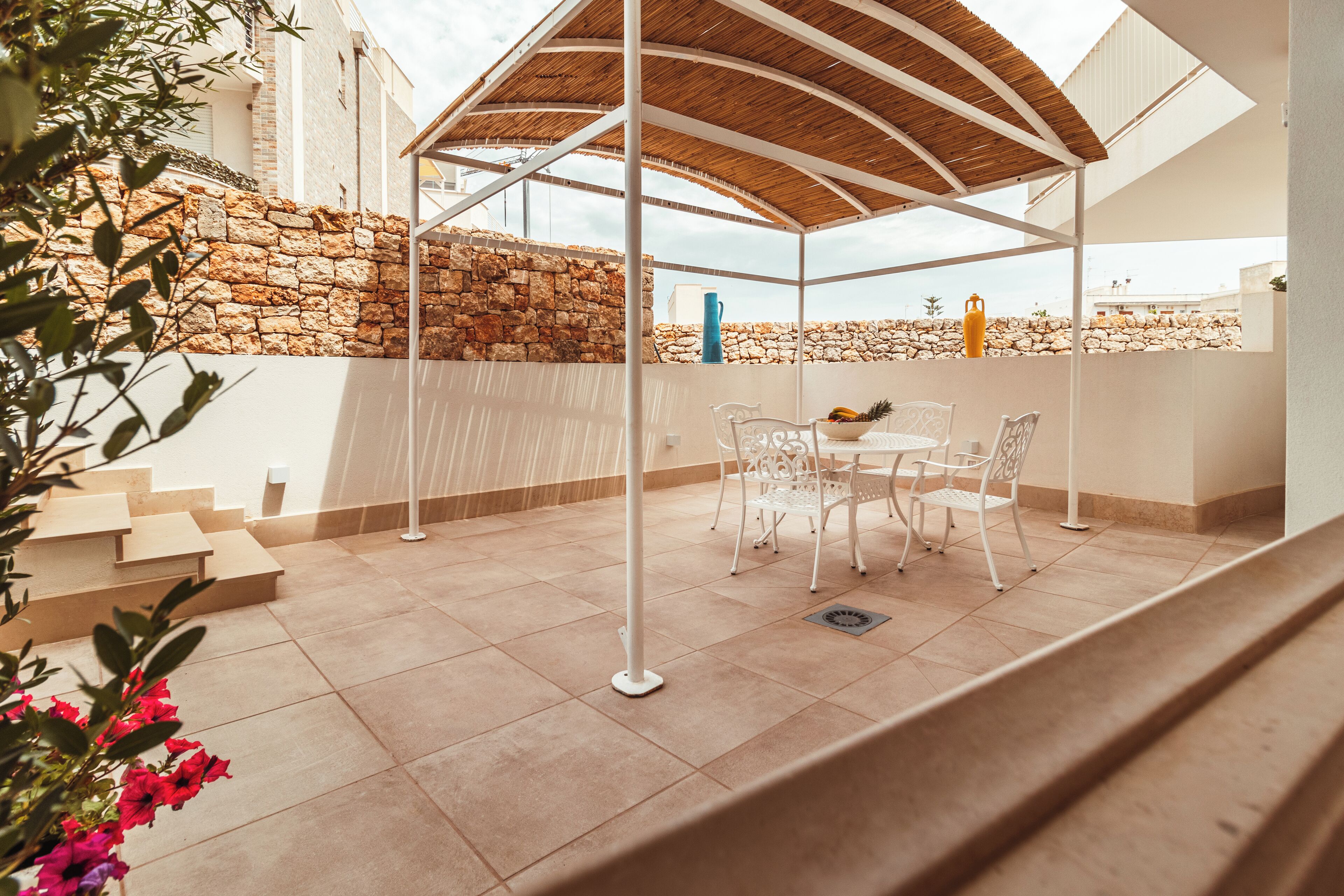 A covered outdoor seating area with a stone wall, white metal chairs, and a table with fruit.