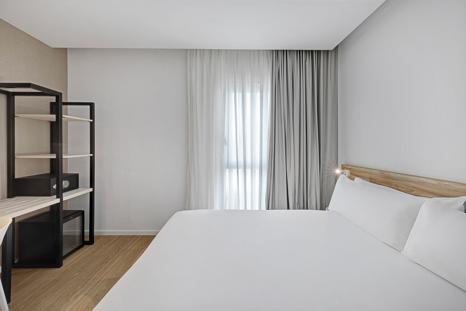 A hotel room with a bed, a desk with shelves, and a window with curtains.