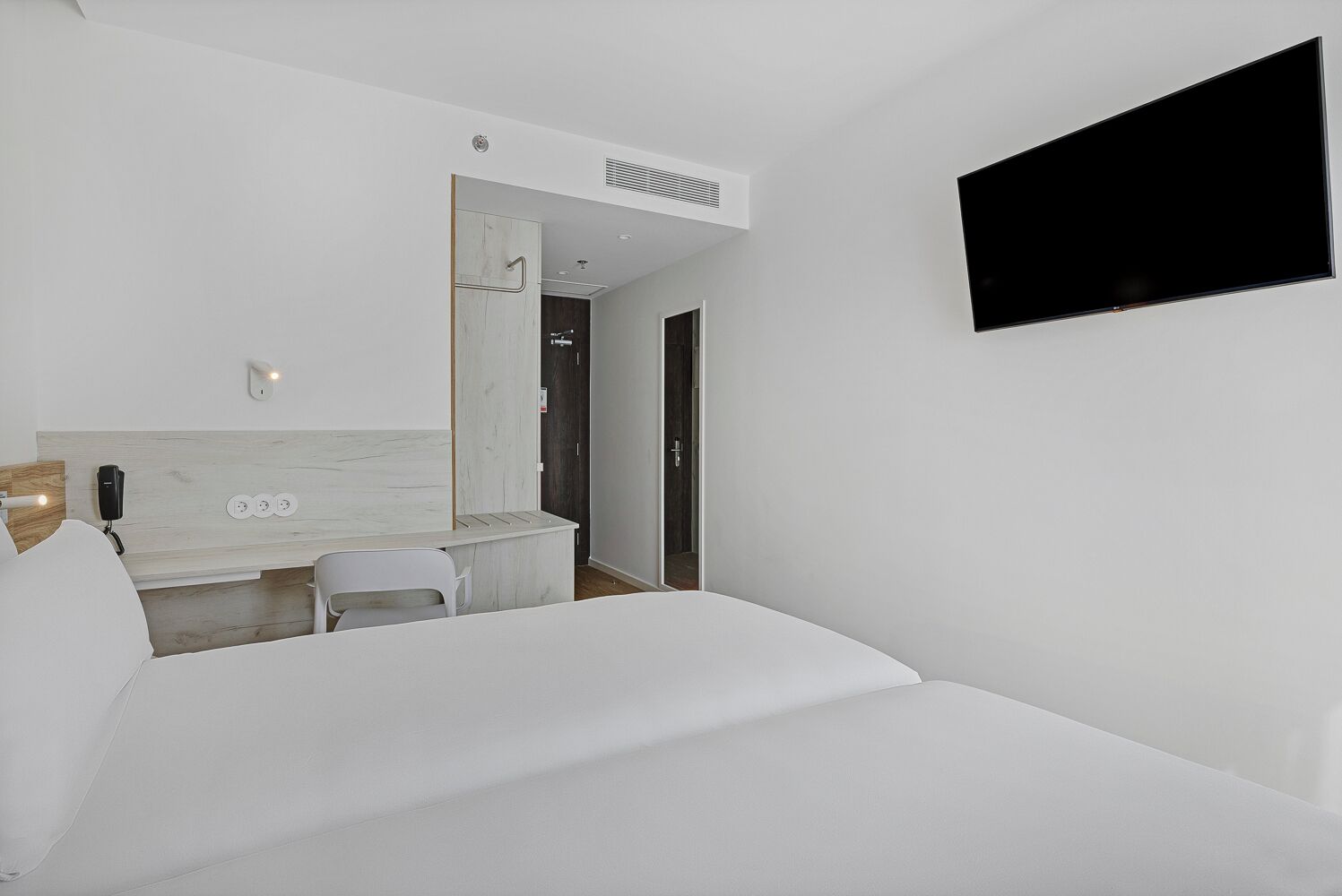 Une chambre d’hôtel moderne équipée d’un lit, d’un bureau avec une chaise, d’un miroir, d’un téléviseur fixé au mur et d’un téléphone sur le bureau.