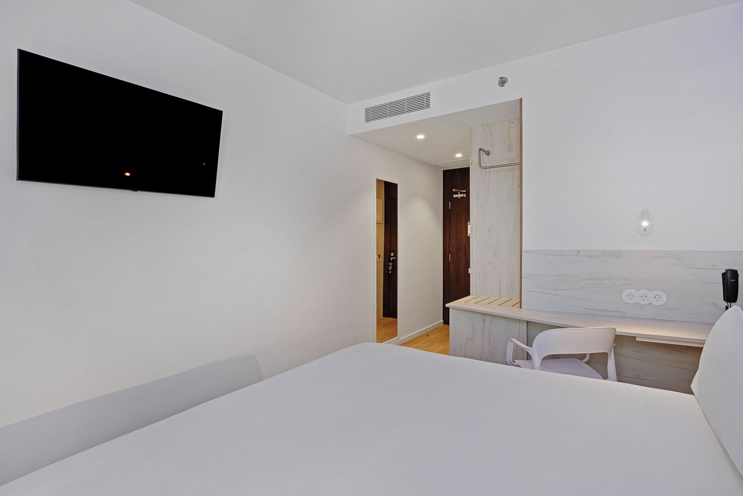 Une chambre d’hôtel moderne, dotée d’un grand lit, d’un bureau avec une chaise, d’un téléviseur fixé au mur et d’une porte en bois.
