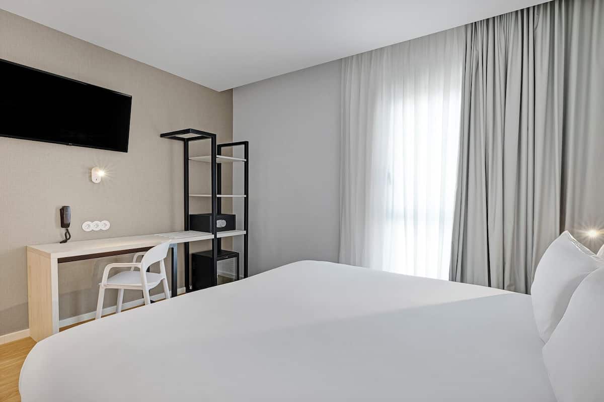 Ein modernes Hotelzimmer mit einem großen Bett, einem Schreibtisch mit Stuhl, einem an der Wand montierten Fernseher und einem Fenster mit Vorhängen.
