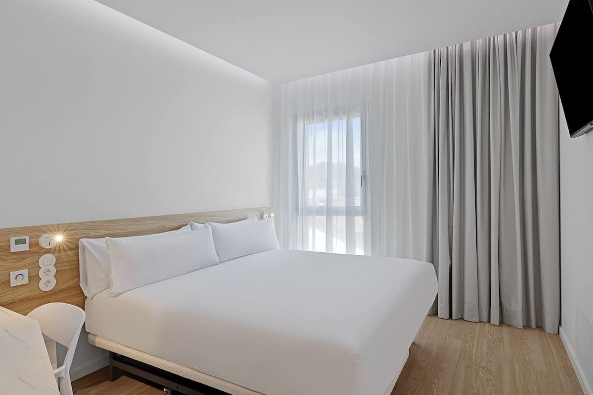 Ein Hotelzimmer mit einem großen Bett, einem Nachttisch mit Bedienfeld, einem Sessel und einem an der Wand befestigten Fernseher.