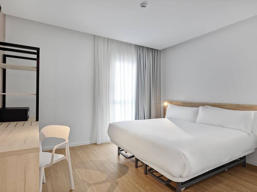 A modern hotel room with a wooden bed frame, white bedding, a desk with a chair, and a large window with curtains.