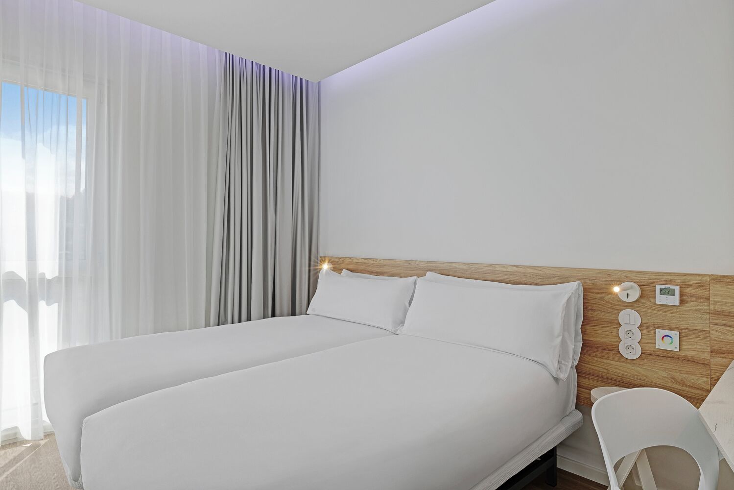 Una habitación de hotel moderna con una cama grande, cabecera de madera y una mesita de noche con lámpara y termostato digital.