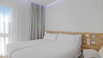 Una habitación de hotel moderna con una cama grande, cabecera de madera y una mesita de noche con lámpara y termostato digital.
