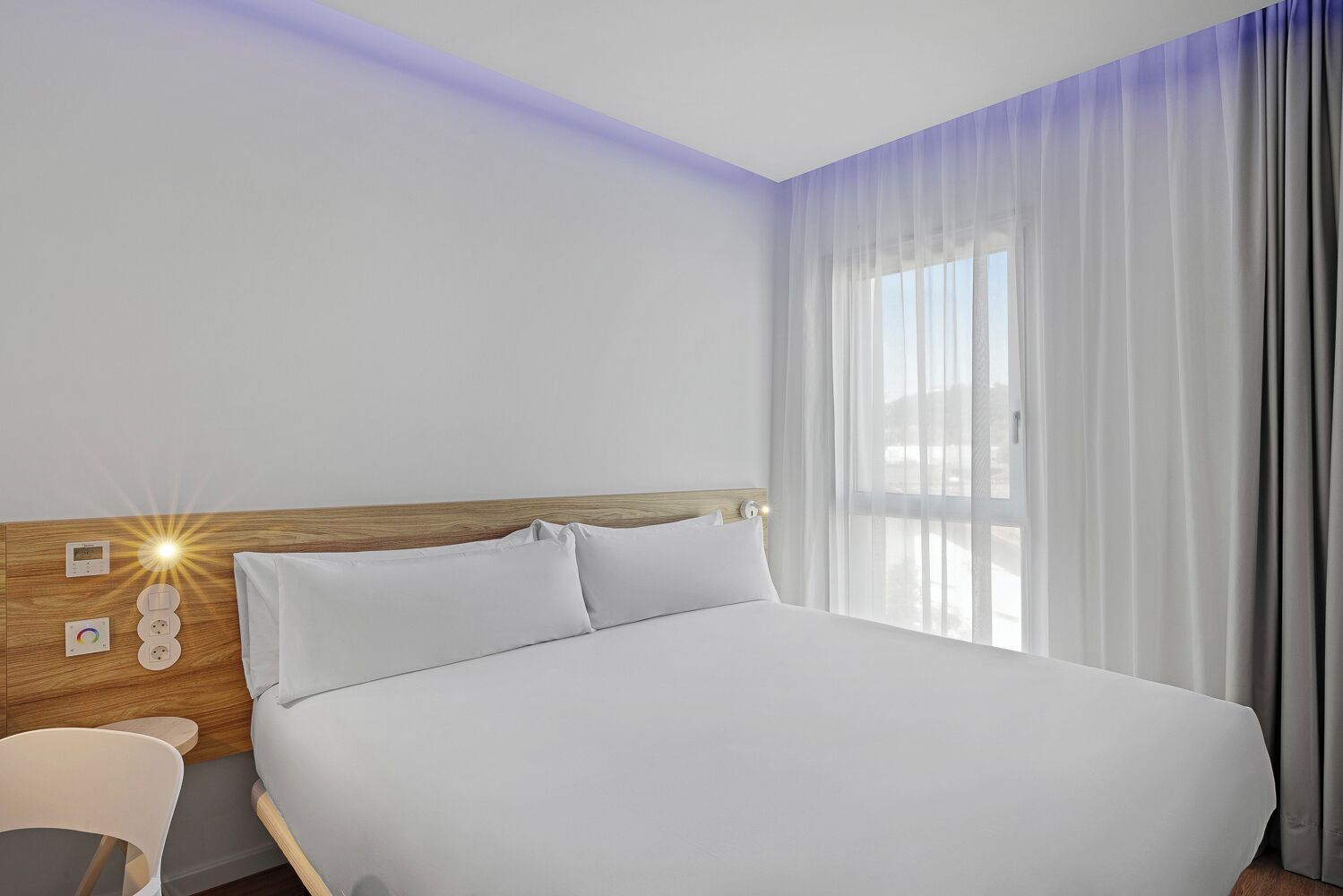 A modern hotel room with a large bed, a wooden headboard, a bedside table, and a window with sheer curtains.
