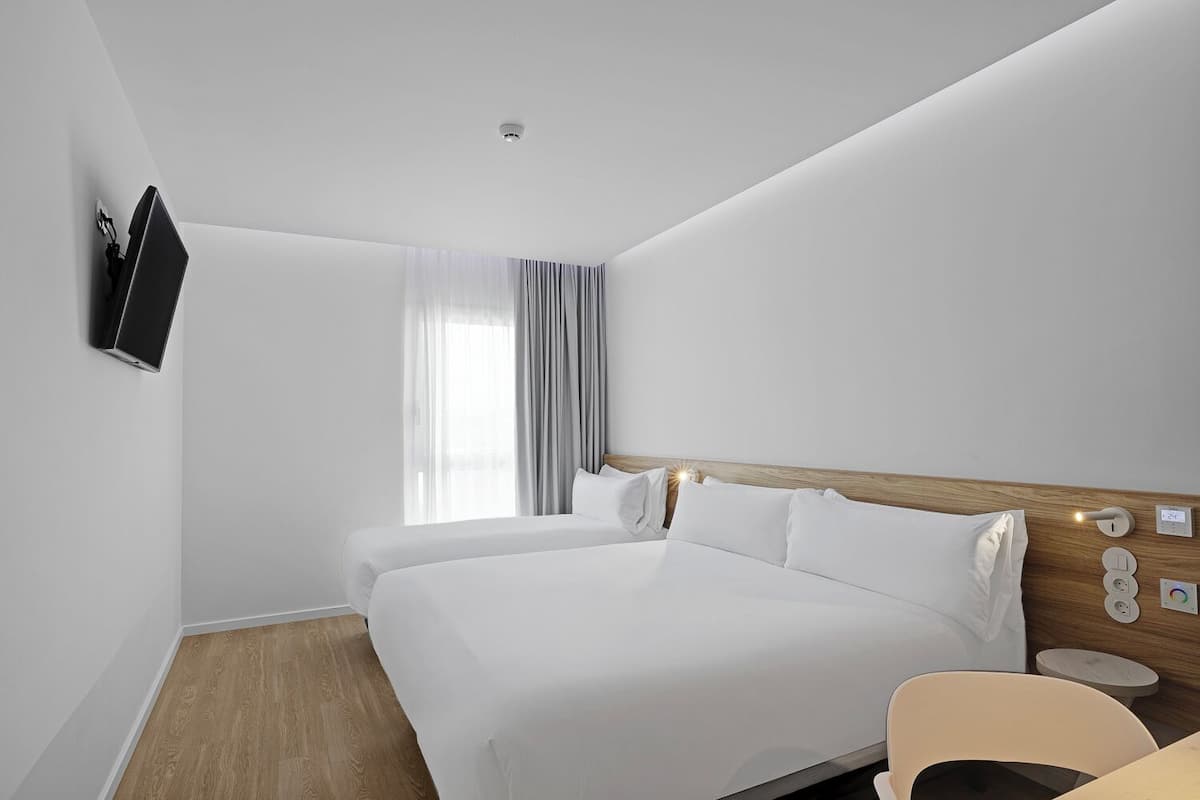 Ein Hotelzimmer mit zwei Betten, einem an der Wand montierten Fernseher, einem Nachttisch mit Lampe und einem Fenster mit Vorhängen.