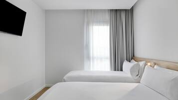 Ein Hotelzimmer mit zwei Betten, einem an der Wand montierten Fernseher und einem mit Vorhängen versehenen Fenster.