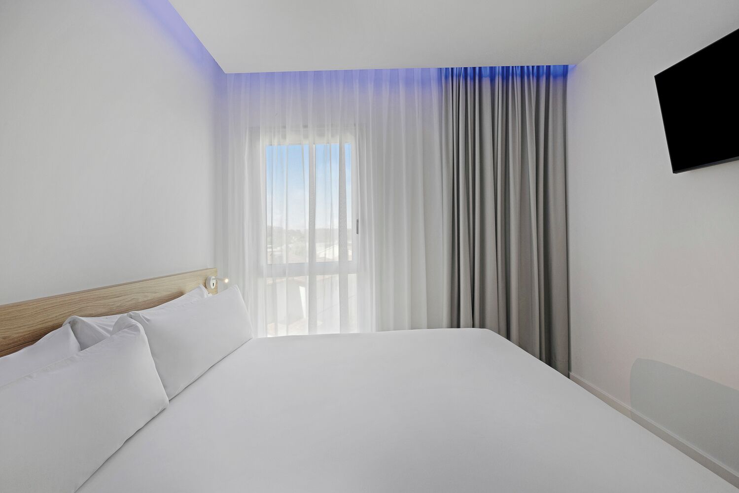 A hotel room with a large bed, a wooden headboard, a window with sheer curtains, and a flat-screen TV mounted on the wall.