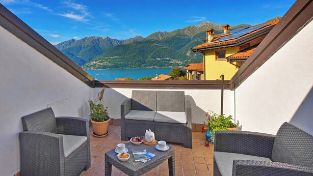 Een balkon met rieten meubilair, een tafel met ontbijtartikelen en uitzicht op bergen en een meer.
