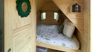 Una pequeña habitación tipo cabaña de madera, con una cama, un estante con libros y una ventana con cortinas.