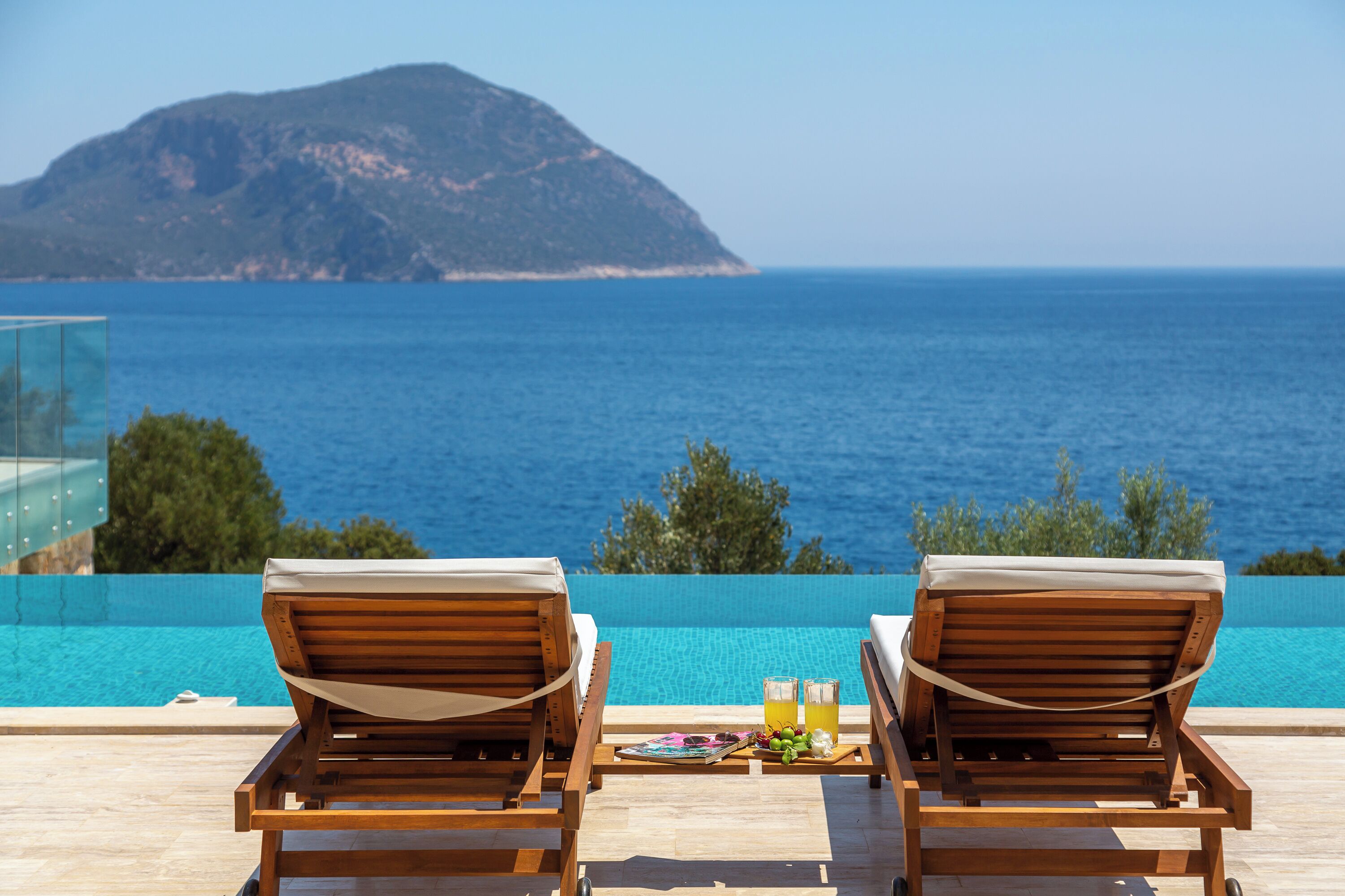 Two wooden lounge chairs with cushions, a tray with a drink and a magazine, and a clear view of the sea and a distant island.