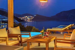 Un área de estar al aire libre con madera, una piscina, sillones reclinables y una mesa con bebidas y flores.