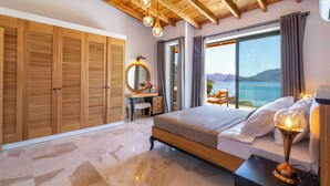 A bedroom with a wooden ceiling, a large bed, a desk with a mirror, and a view of the sea.