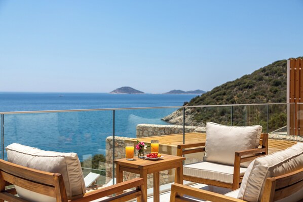 Un balcón con muebles de madera, una mesa con bebidas y fruta, y una vista panorámica del mar y las colinas.