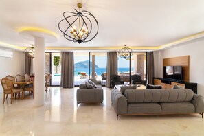 A spacious living area with a large sofa, a dining table, and chairs. There is a TV mounted on the wall and a chandelier hanging from the ceiling.