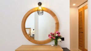 A round mirror with a wooden frame, a wooden desk with a black top, a vase with flowers, and a wooden door with louvered panels.