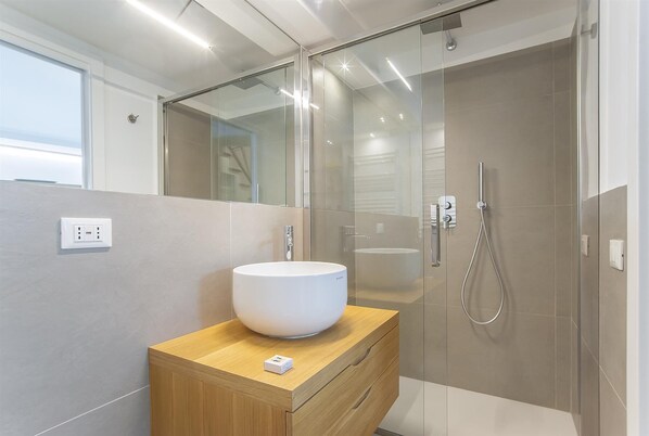 Un bagno moderno con un ampio lavandino bianco, un mobile contenitore in legno, una cabina doccia in vetro e una presa elettrica a vista.