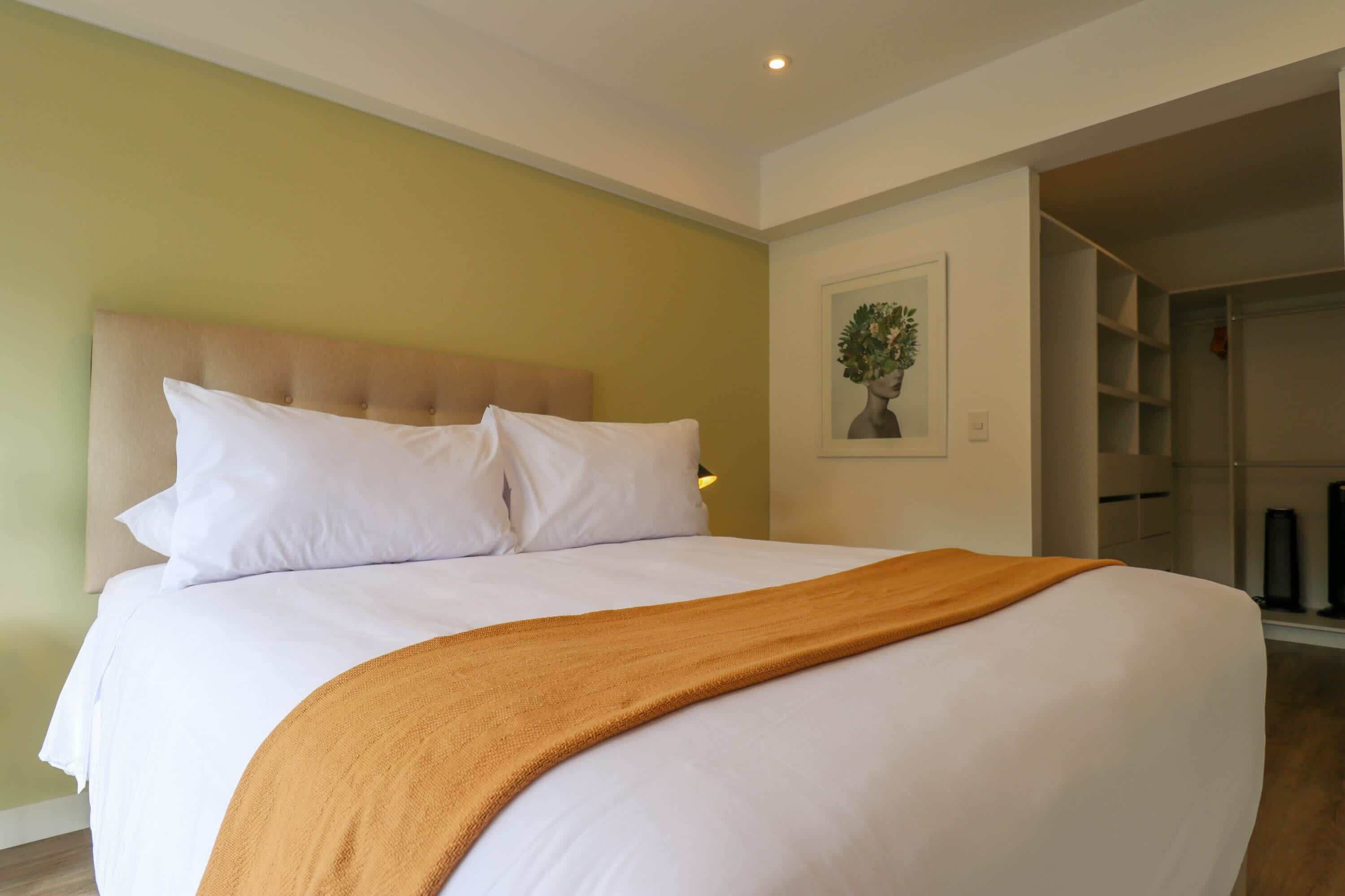 A neatly made bed with white linens and a brown throw blanket, a beige headboard, and a framed picture on the wall.