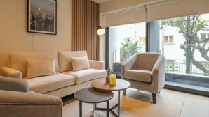 Ein modernes Wohnzimmer mit einer Couch, einem Sessel und einem Couchtisch. Ein gerahmtes Stadtbild hängt an der Wand, und ein Fenster bietet Blick auf Gebäude und Bäume.