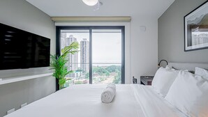 A modern hotel room with a large bed, a flat-screen TV mounted on the wall, a nightstand with a lamp, and a view of the city through a large window. - w* | Minimalist 1BR w/ Balcony in San Francisco (Panamá)