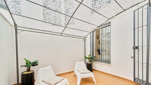Ein überdachter Balkon mit zwei weißen Stühlen, Topfpflanzen und einem Fenster mit Gitterstäben.