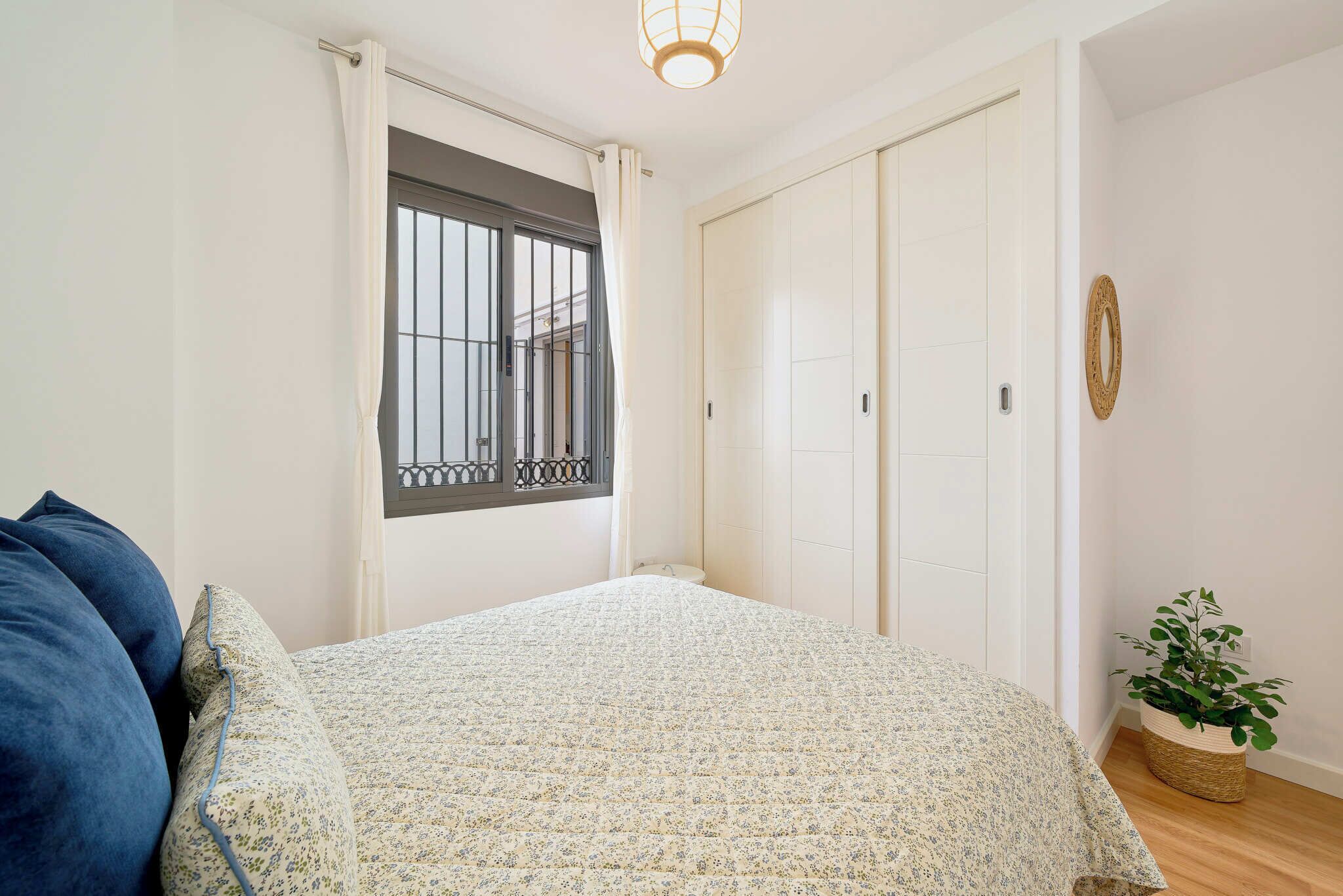 A bedroom with a bed, a window with bars, a white wardrobe, and a potted plant.